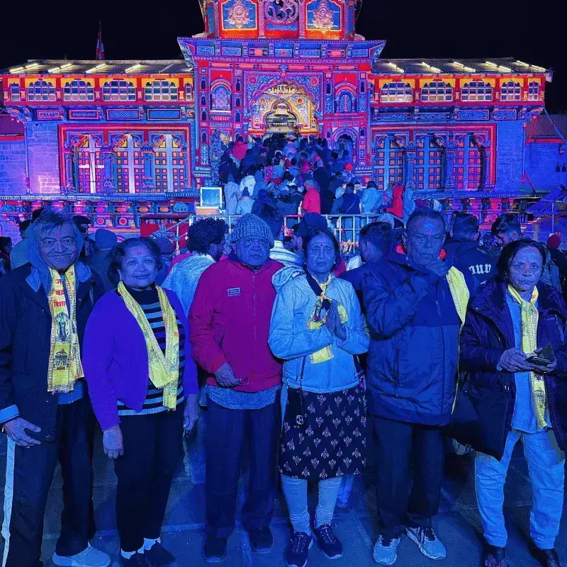 Happy elderly clients posing in front of the brightly lit Badrinath temple during their Chardham tour with us (chardham package).